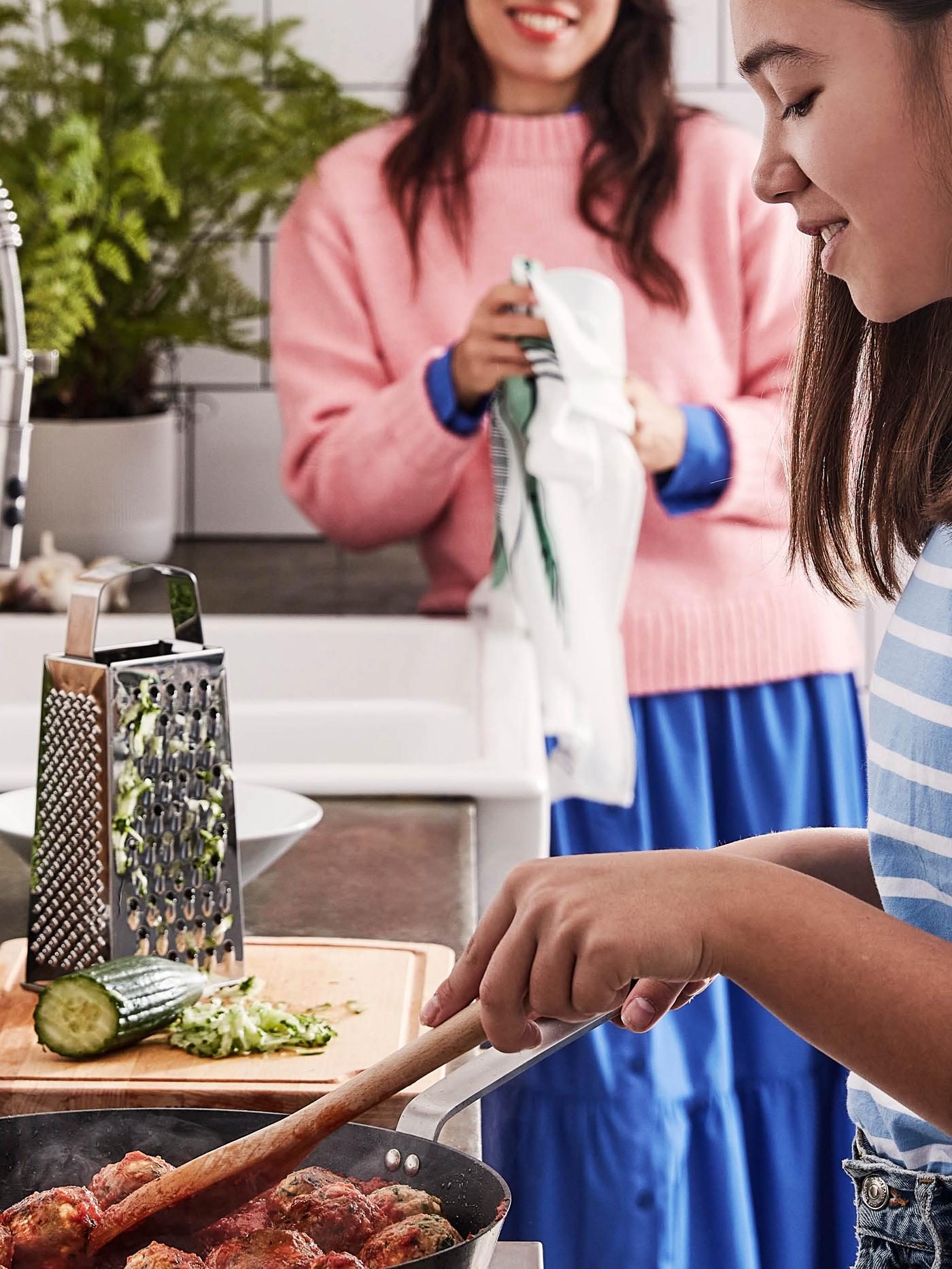Dívka v pruhovaném tričku míchá v pánvi VARDAGEN rajčatovou omáčku s kuličkami z bezmasé směsi VÄRLDSKLOK, vedle na prkénku PROPPMÄTT leží struhadlo IDEALISK s půlkou nastrouhané okurky.