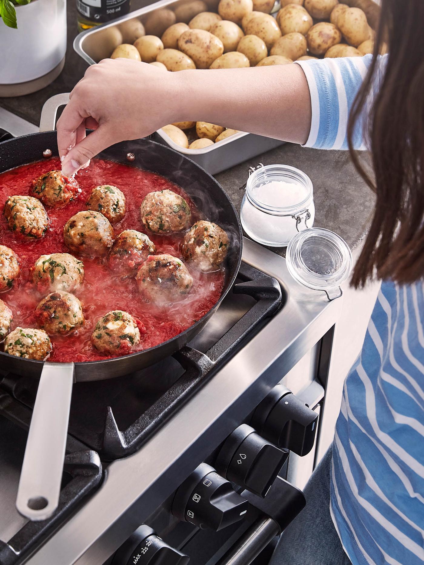 Dívka v pruhovaném tričku solí rajčatovou omáčku s kuličkami z bezmasé směsi VÄRLDSKLOK v pánvi VARDAGEN.