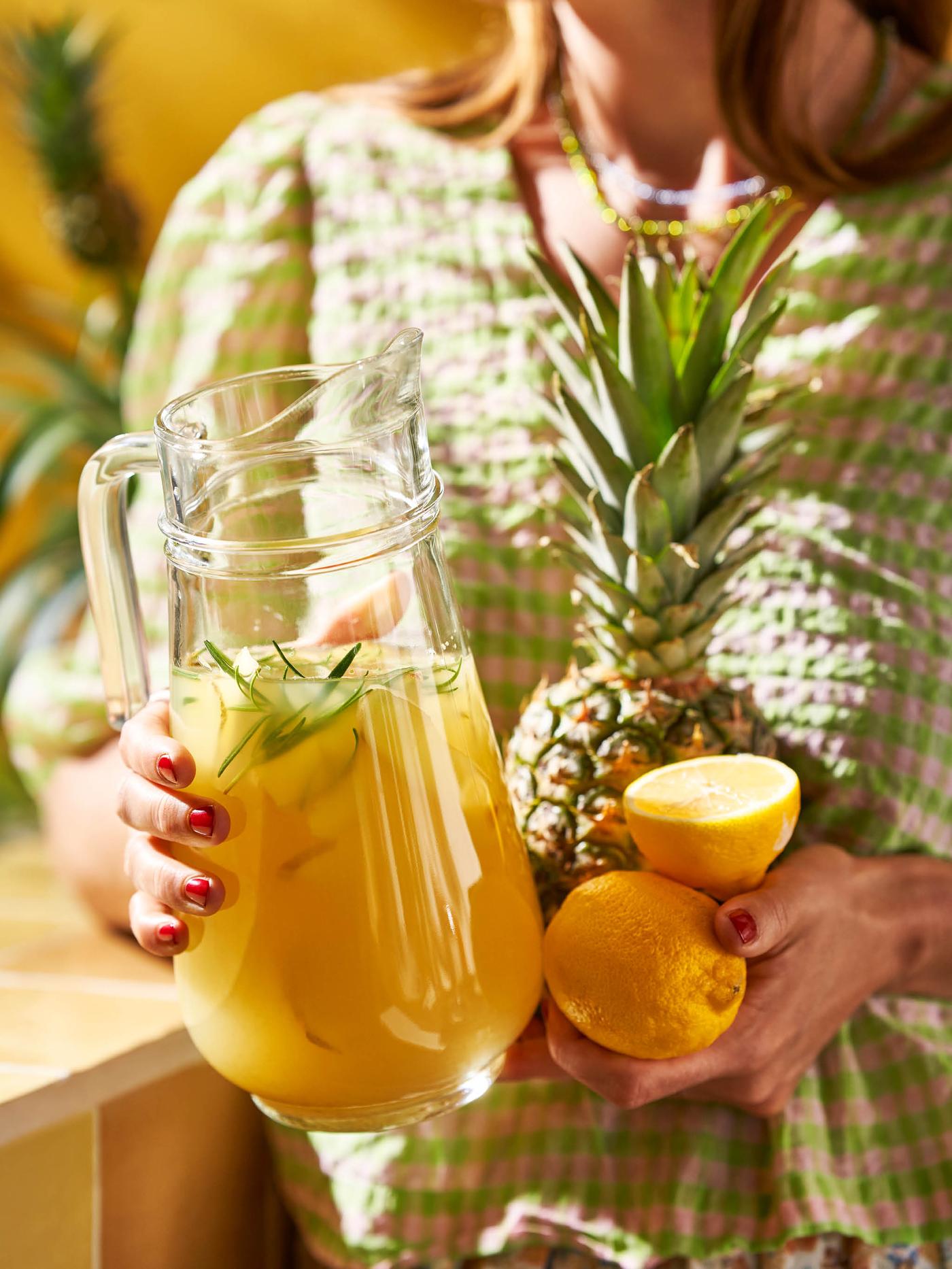 Dívka v světlezelno-bílých šatech drží v jedné ruce džbán TILLBRINGARE s ovocným nápojem, ve druhé ruce ananas a dva citrony.