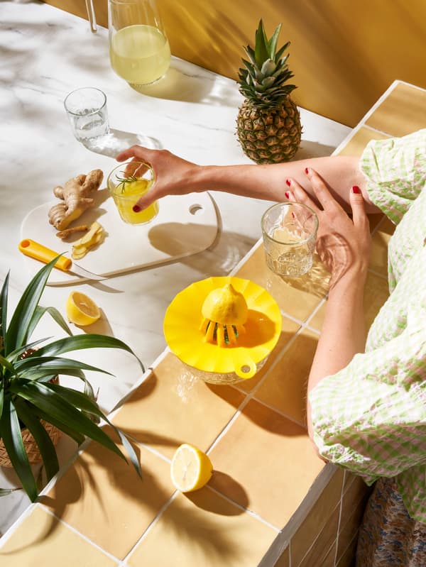 Dívka v světlezelno-bílých šatech odšťavňuje citrony pomocí odšťavnovače UPPFYLLD a staví sklenici citronové šťávy na prkénko vedle kousku čerstvého zázvoru a nože UPPFYLLD.