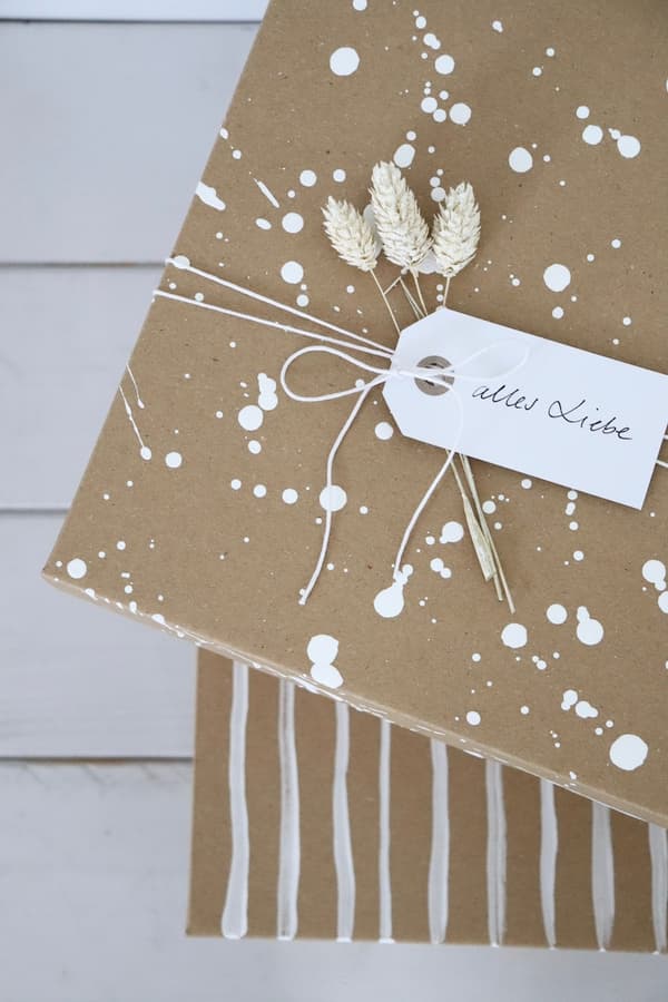 DIY Geschenkkarton: Zwei VATTENTRÅG Kasten aus Pappkarton in der Farbe braun, aufeinandergestapelt. Beide sind mit weißer Farbe bemalt. Der untere mit einem Linienmuster, der obere mit Tupfen. Eine Schleife aus Naturfaden, Trockenblumen und ein weißer Geschenkanhänger verzieren den oberen Kasten. 