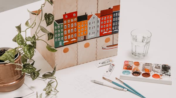 DIY Idee: KNUFF Zeitschriftensammler mit buntem Häusermotiv bemalen. Daneben liegen alle Utensilien, wie Bleistifte, Gouache-Farben, Pinsel, ein Wasserglas und Klebetape.