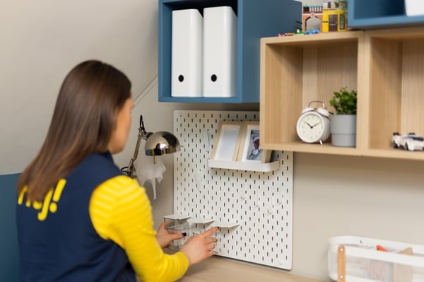 Дизайнер IKEA расставляет предметы на детском рабочем столе, корректируя аксессуары на перфорированной панели и полках для создания организованного и функционального пространства.