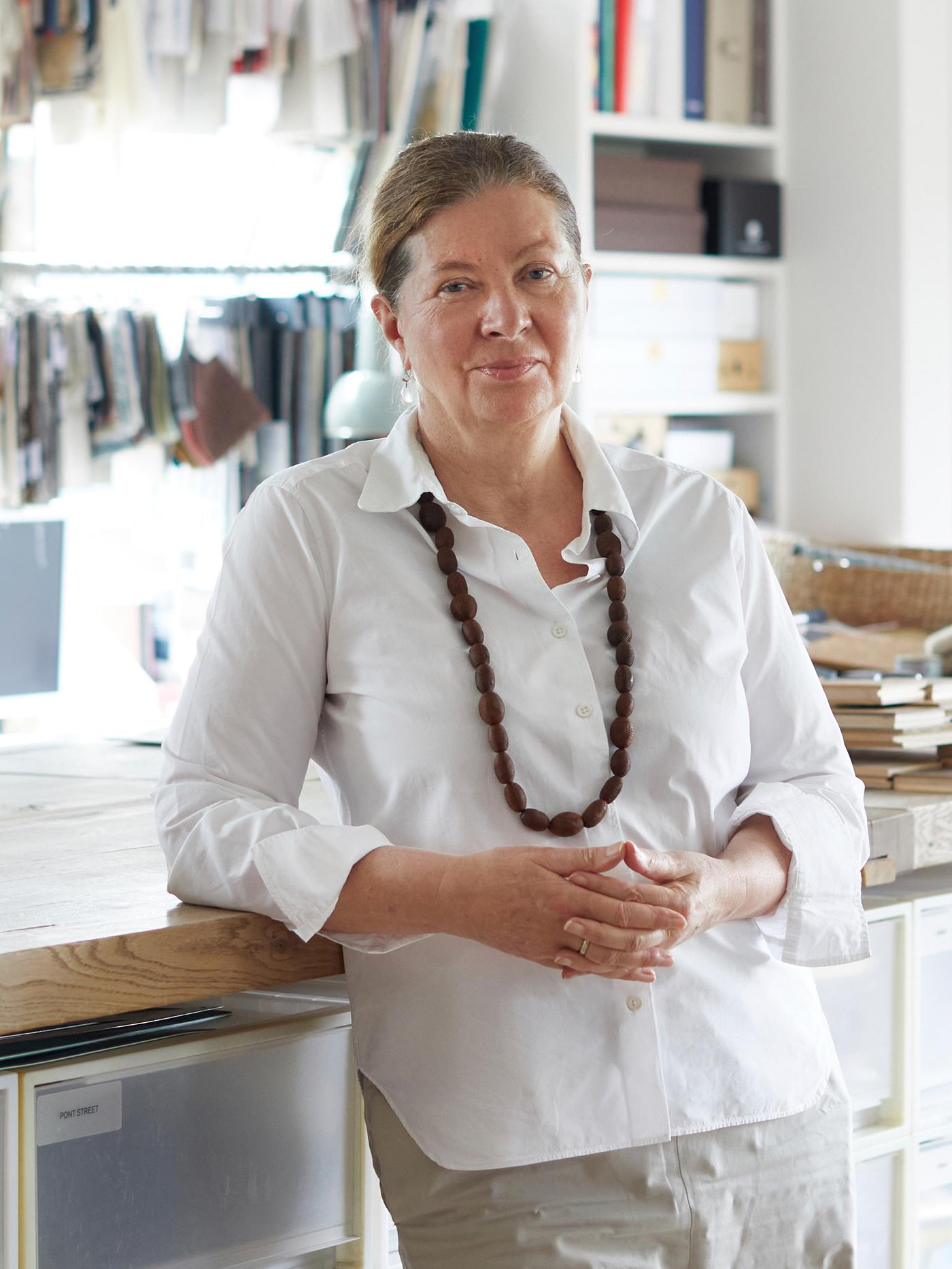 Dizajnerka Ilse Crawford, koja nosi belu košulju i glomaznu ogrlicu, naslonjena je na radnu ploču u svom studiju.