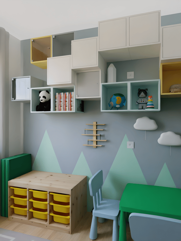 Dječja soba s zidnim policama, plišanim igračkama, knjigama i oblačićima. Stol s plastičnim ladicama i dvije stolice. Zid ukrašen nacrtanim planinama.