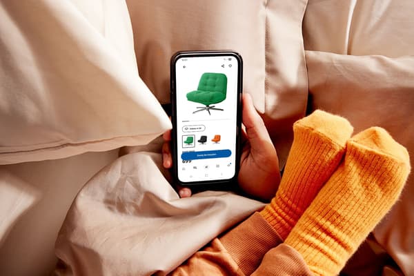 Dłoń trzyma smartfon z uruchomioną aplikacją IKEA na tle pościeli i obok stóp dziecka w pomarańczowych skarpetkach.