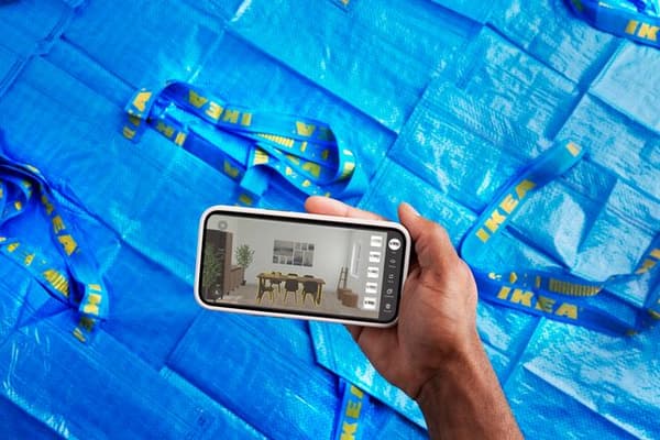 Dłoń trzymająca smartfon z uruchomioną aplikacją IKEA. Tło stanowią niebieskie torby FRAKTA.