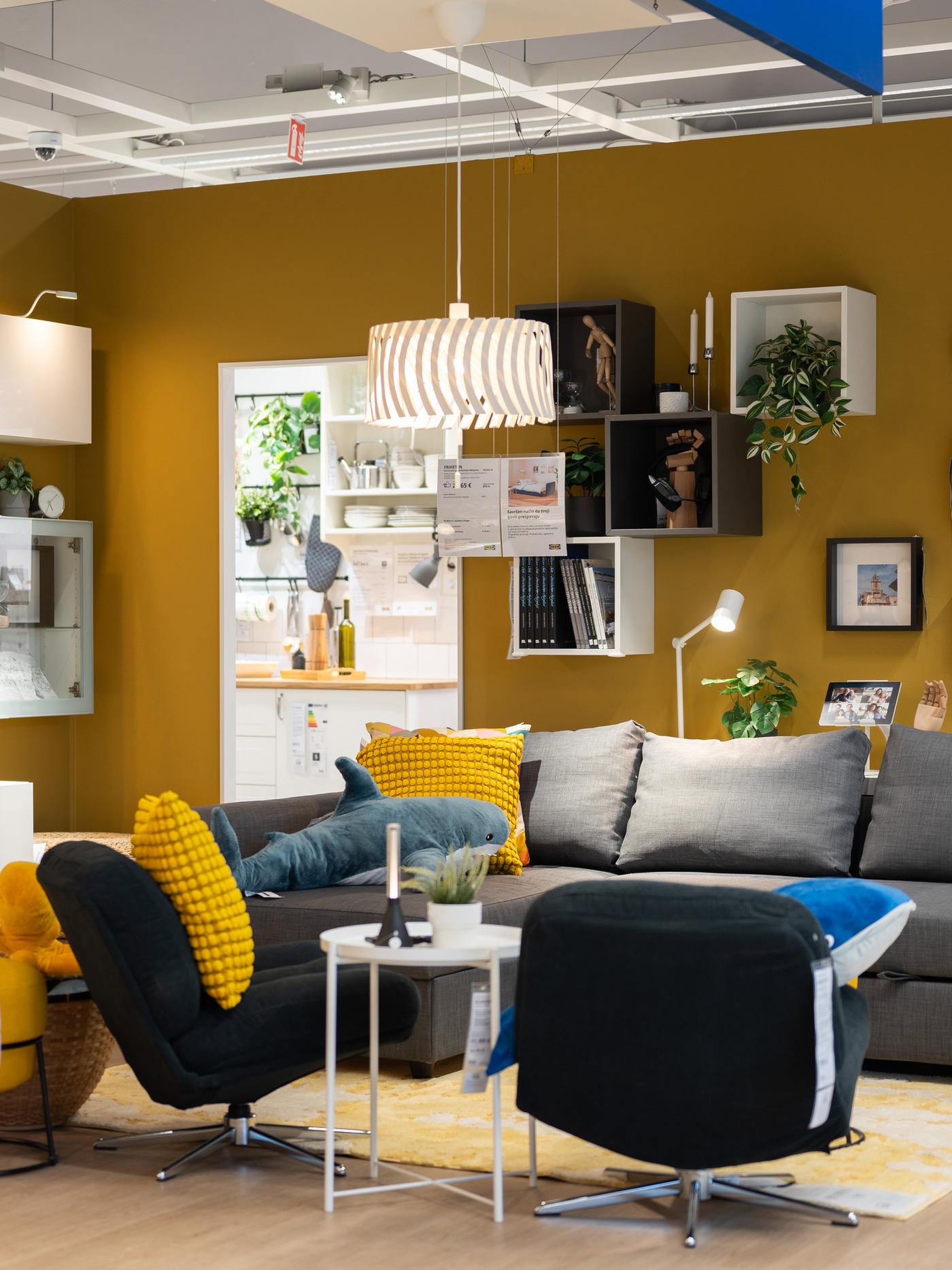 Dnevni boravak s modernim namještajem, uključujući sivu sofu, crne fotelje i žute jastuke, te viseću svjetiljku i police na zidu.