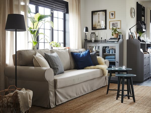 Dnevni boravak sa sofom na razvlačenje u svetlobež boji, tepih od jute, sivi ormarić sa staklenim vratima i dva seta stočića u tamnim, plavim i zelenim nijansama.