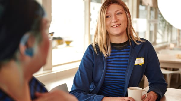 Doi angajați IKEA, îmbrăcați în uniformă de lucru, savurează o cafea împreună în restaurantul pentru angajați.