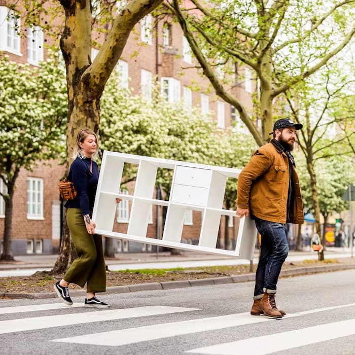 Doi oameni cară împreună o etajeră albă IKEA peste o trecere de pietoni, într-un cartier verde cu copaci și clădiri de cărămidă, sugerând mobilă ușor de transportat acasă.