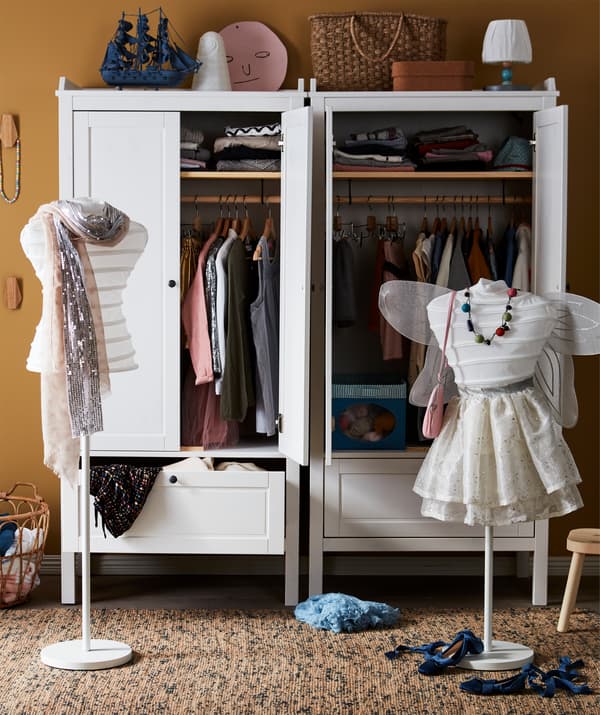 Dois cabides brancos parcialmente cobertos com roupa a imitar troncos de manequim, colocados em frente a dois roupeiros com roupa.