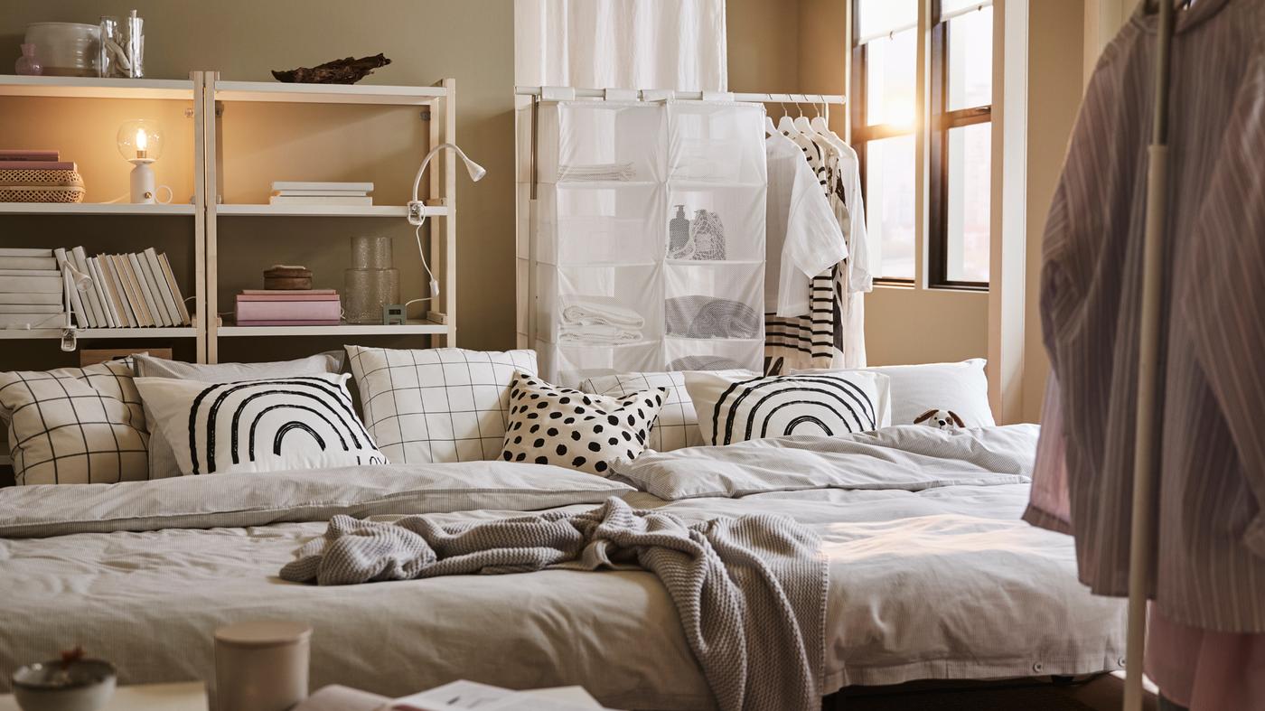 Dois sofás-cama LYCKSELE LÖVÅS com roupa de cama em cinzento e almofadas com padrões, estantes em branco/choupo e suportes para cabides em branco.