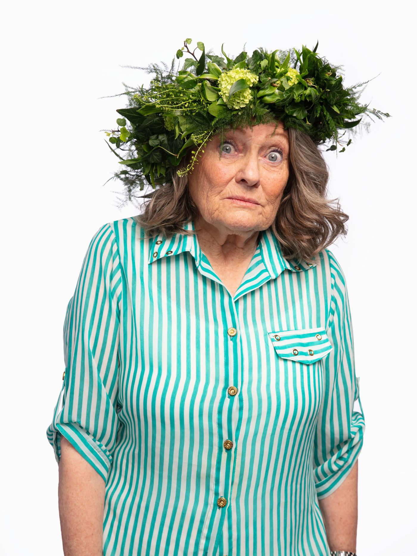 Dona gran amb expressió sorpresa, camisa de ratlles verdes i blanques i corona de fulles al cap.
