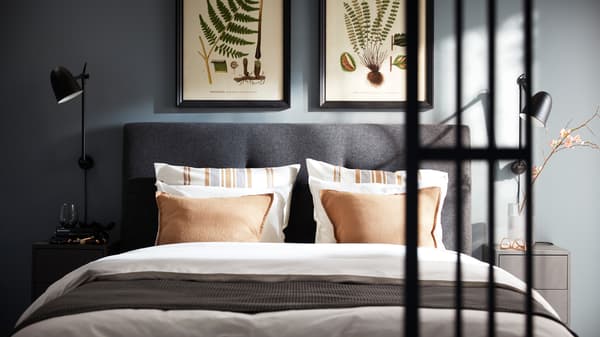Donkergrijs IDANÄS bed met wit ÄNGSLILJA bedlinnen, donkergrijze VÅRELD sprei en zwarte SKURUP wandlampen aan weerszijden.