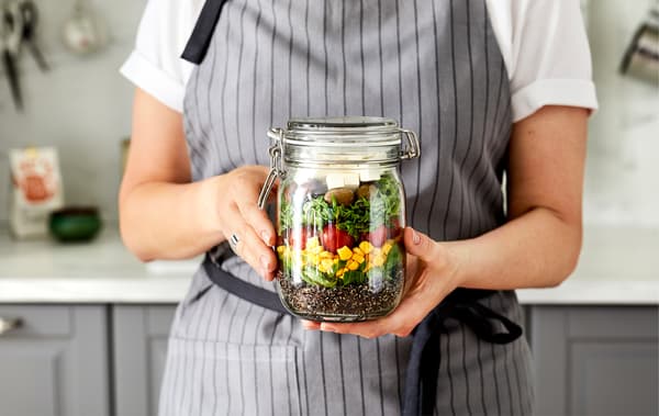 Donna con grembiule in cucina regge un barattolo contenente un’insalata a strati colorati - IKEA