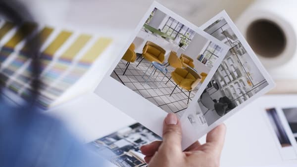 Donna di spalle che tiene in mano due fotografie che ritraggono arredi di un moderno spazio d'ufficio.