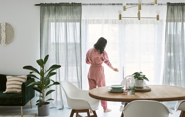 Donna in tuta rosa sistema le tende davanti a una portafinestra in una sala da pranzo con un tavolo in legno e sedie bianche - IKEA