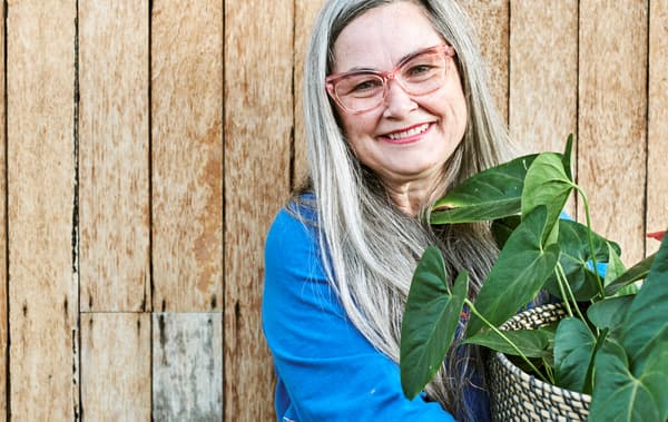 Donna sorridente con lunghi capelli grigi, occhiali dalla montatura rosa e una maglia blu tiene in mano un vaso intrecciato con dentro una pianta a foglie larghe.