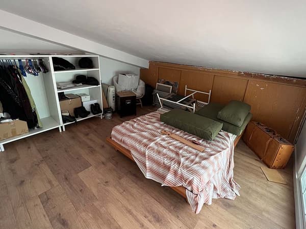 Dormitori sota teulada amb un llit, un armari, roba penjada en penja-robes, mobles amuntegats, un cobrellit i coixins.