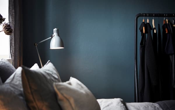 Dormitorio coa parede de cor azul escuro e verde. Hai unha cómoda cama, unha lámpada branca e un colgadoiro.