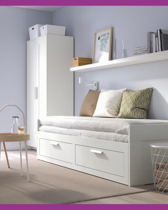 Dormitorio con cama blanca con cajones inferiores, cojines decorativos y repisa con objetos sobre la pared.