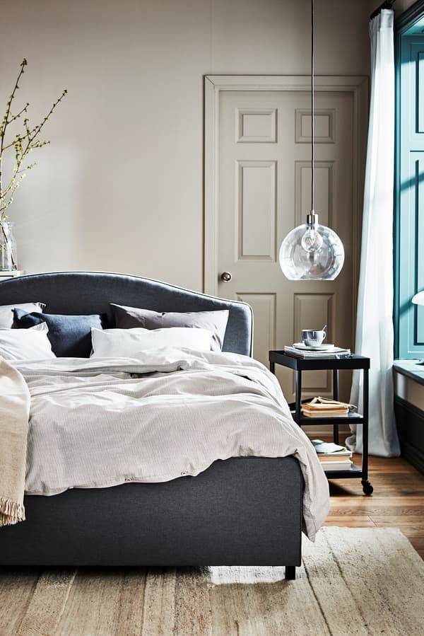 Dormitorio con una cama de base gris y un velador al lado derecho con una taza de café.