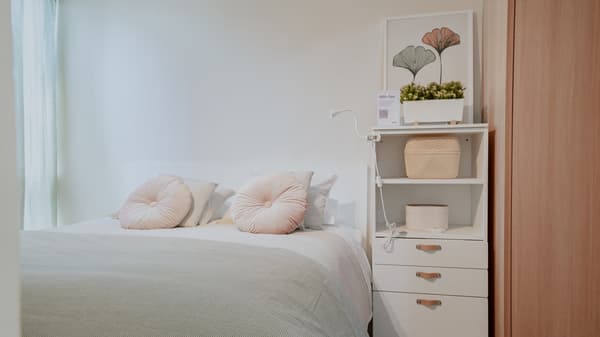 Dormitorio principal con cama doble con cubrecama gris, sabanas blancas y cojines rosados redondos. Junto a ella hay un estante blanco con cajones.