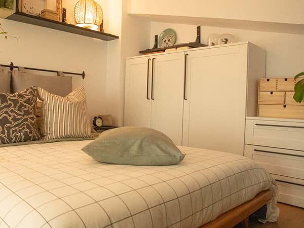 Dormitorio tras ser reformado con cama con colcha y cojines, armario, cómoda, baldas, cajonera de madera, lámpara y adornos.