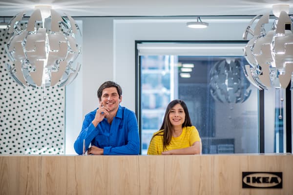 Dos colaboradores, en polera azul y amarilla, sonriendo. Se encuentran detrás del mesón de recepción de las oficinas IKEA.