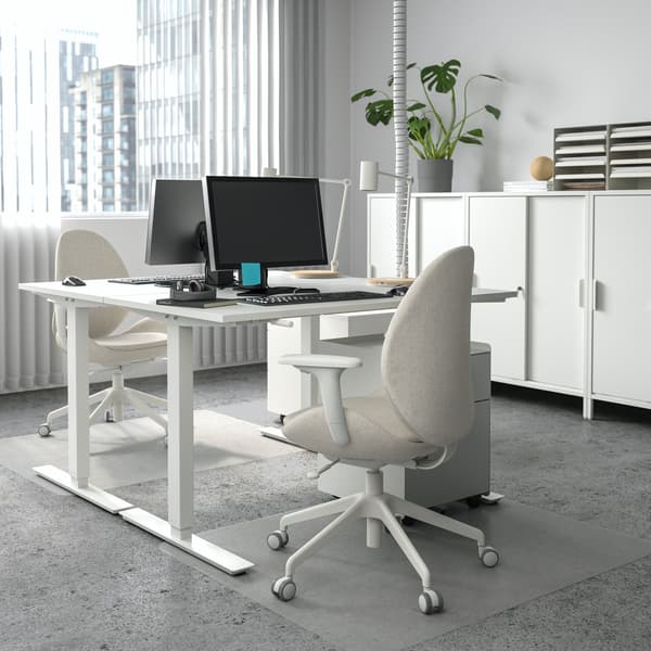 Dos escritorios de altura ajustable blancos con monitores y teclados, frente a sillas de escritorio de color blanco crema, en una oficina con piso de hormigón y un ventanal que muestra un entorno corporativo. 