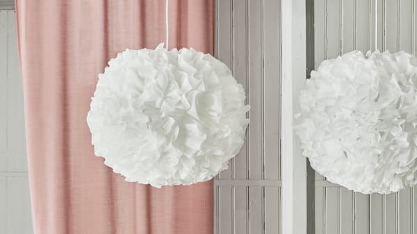 Dos lámparas de techo VINDKAST apagadas con forma de pompón blanco y mullido colgadas frente a una cortina rosa.
