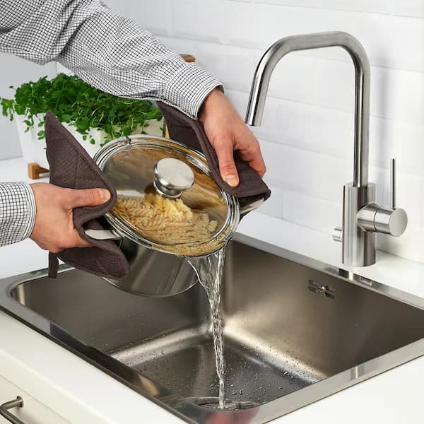 Dos manos inclinando una olla HEMKOMST de acero inoxidable con tapa de vidrio sobre un lavaplatos, para colar el agua con ayuda de la misma tapa. 