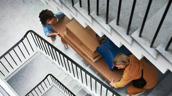 Dos personas suben varias cajas grandes de IKEA y una bolsa azul de IKEA por las escaleras grises de un edificio de apartamentos.
