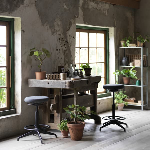 Dos soportes activos de pie/sentado LIDKULLEN en color gris oscuro junto a un espacio de trabajo rodeado de plantas.