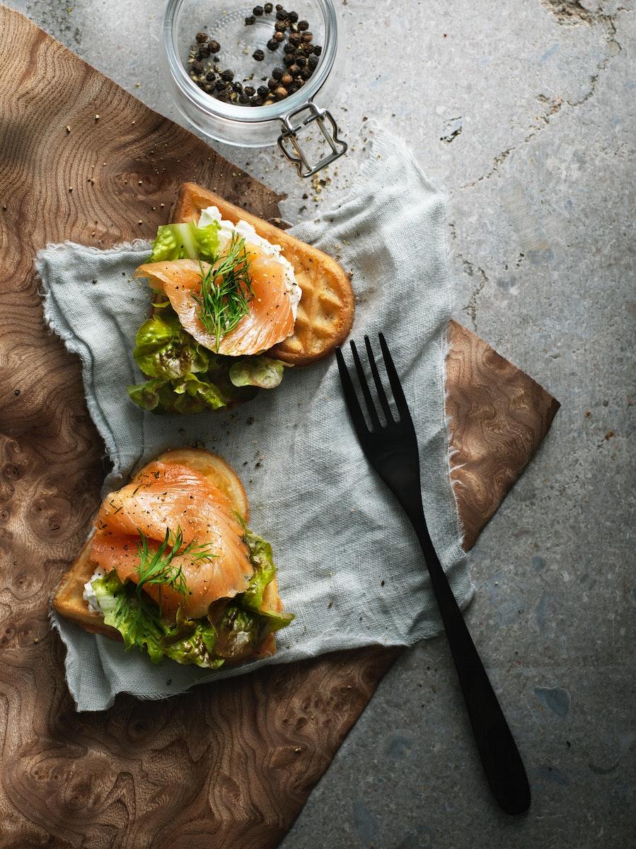 Dos waffles VÅFFLOR en forma de corazón con salmón, queso crema, eneldo y lechuga sobre un paño gris y junto a un tenedor negro. 