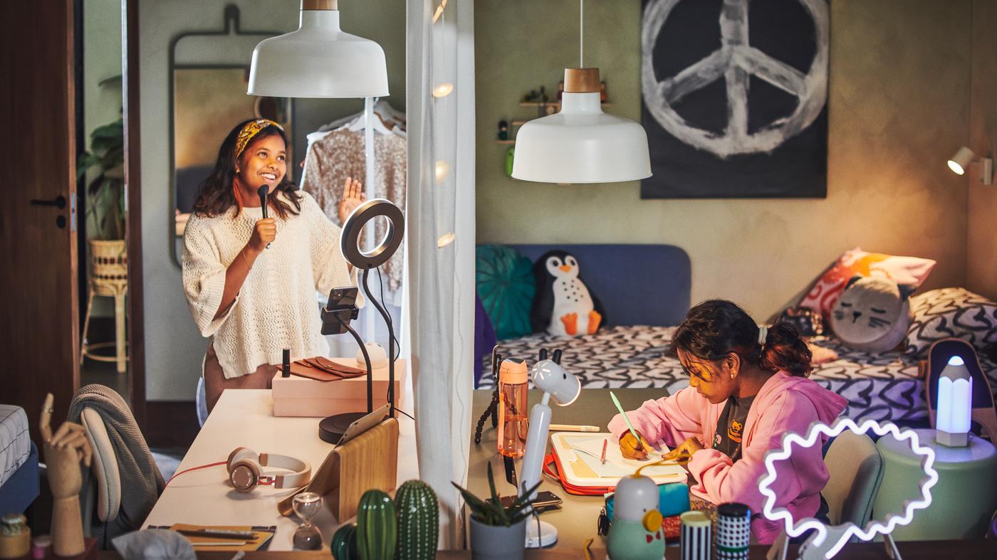 Două adolescente la birouri ALEX așezate față în față. Una studiază, iar cealaltă înregistrează în fața lămpii LÅNESPELARE.