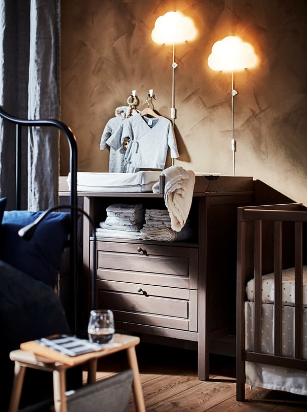 Două aplice UPPLYST cu LED în formă de nor deasupra unei mese de înfășat SUNDVIK maro cu două sertare, lângă un pătuț.