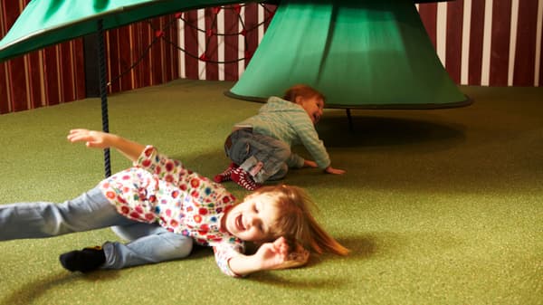 Două copii se joacă pe un covor verde sub o structură mare în formă de ciupercă; unul poartă bluză florală și pantaloni albaștri, celălalt cămașă în carouri și blugi. Fețele sunt blurate.