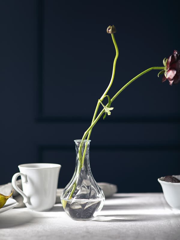 Două flori cu tulpini lungi și așezate într-o vază de sticlă VILJESTARK mică și scurtă pe o față de masă albă.