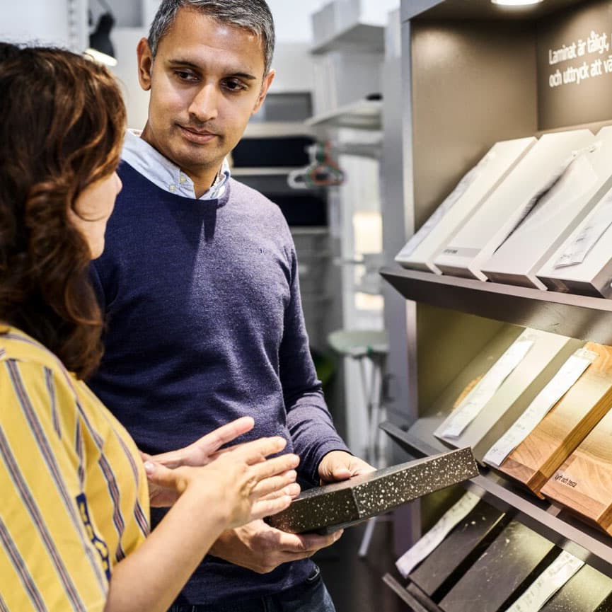 Două persoane discută și examinează mostre de materiale laminate într-un magazin de bricolaj. Persoana din stânga poartă o bluză în dungi galbene, iar persoana din dreapta poartă un pulover albastru.