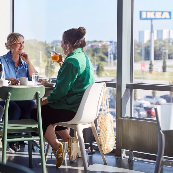 Două persoane stau la o masă într-o cafenea, discutând și bând băuturi. În fundal, prin ferestrele mari, se vede un semn IKEA și o parcare.