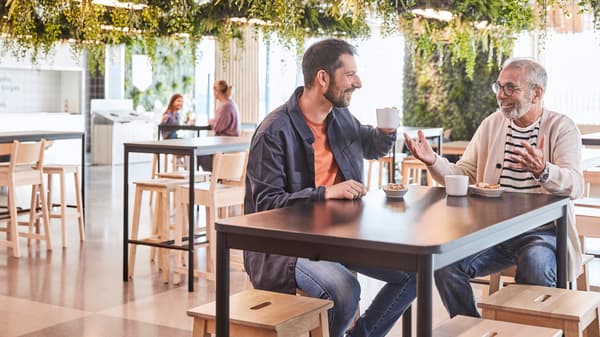 Două persoane stau la o masă într-o cafenea modernă, discutând și ținând cești de cafea. În fundal, alte două persoane sunt așezate la o altă masă. Decorul include multe plante suspendate și mobilier din lemn deschis la culoare.