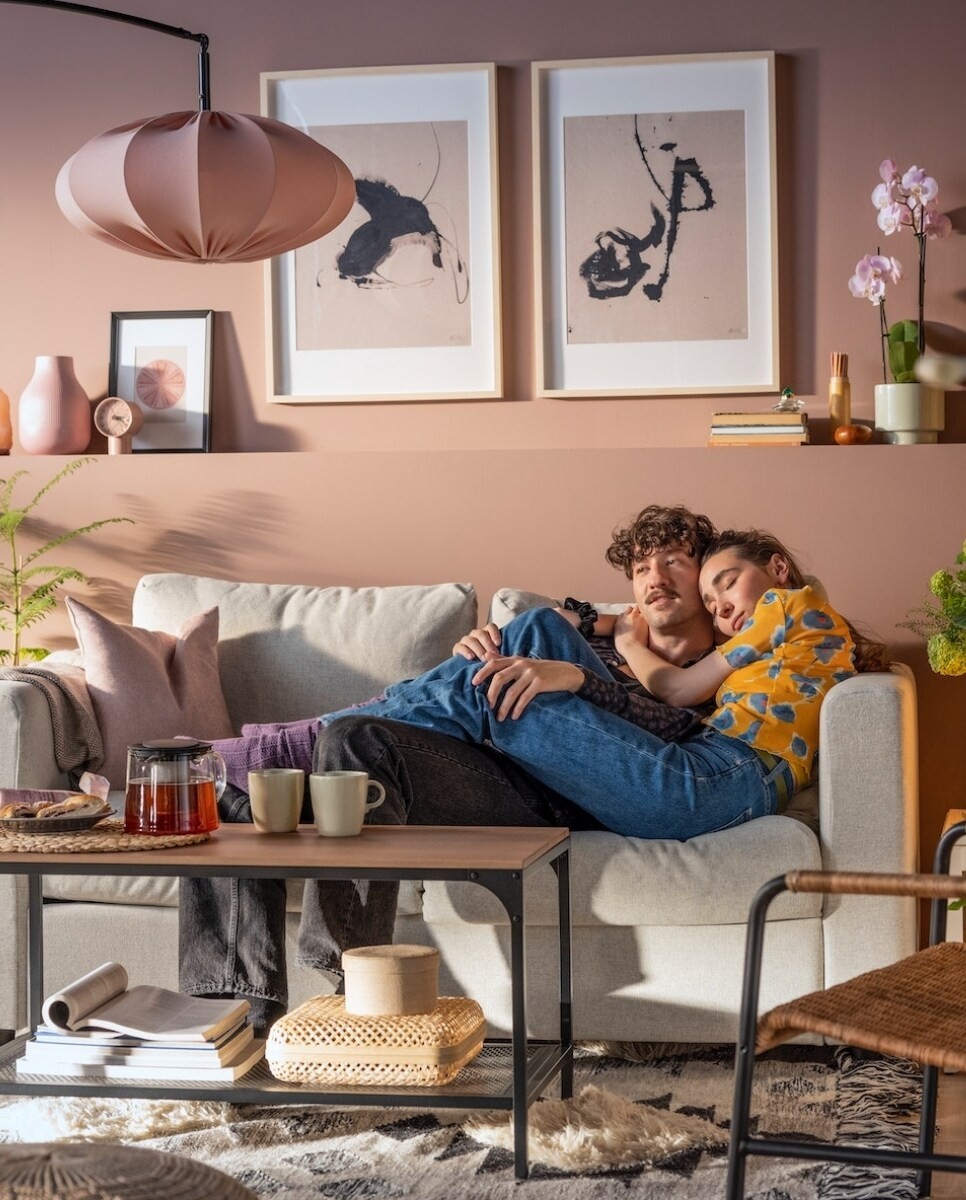 Două persoane stau pe o canapea într-o cameră de zi decorată modern. Pe peretele din spatele lor sunt două tablouri abstracte. În fața canapelei se află o măsuță de cafea cu cești, un ceainic și alte obiecte decorative.