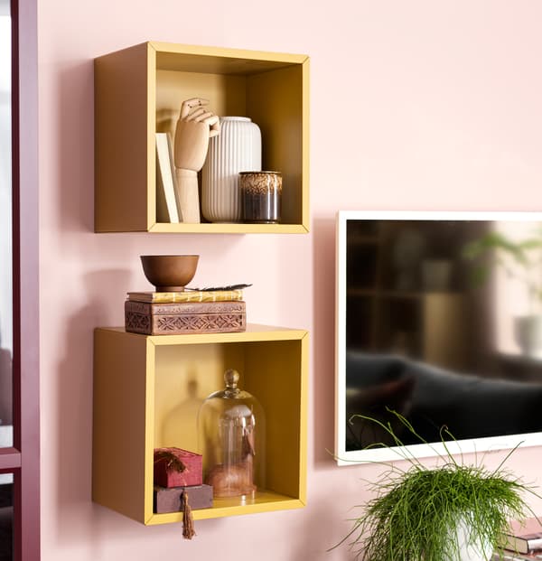 Două unități de depozitare deschisă EKET galbene, suspendate pe perete, cu compoziții din vaze ceramice, forme din lemn, cărți și cutii decorative.