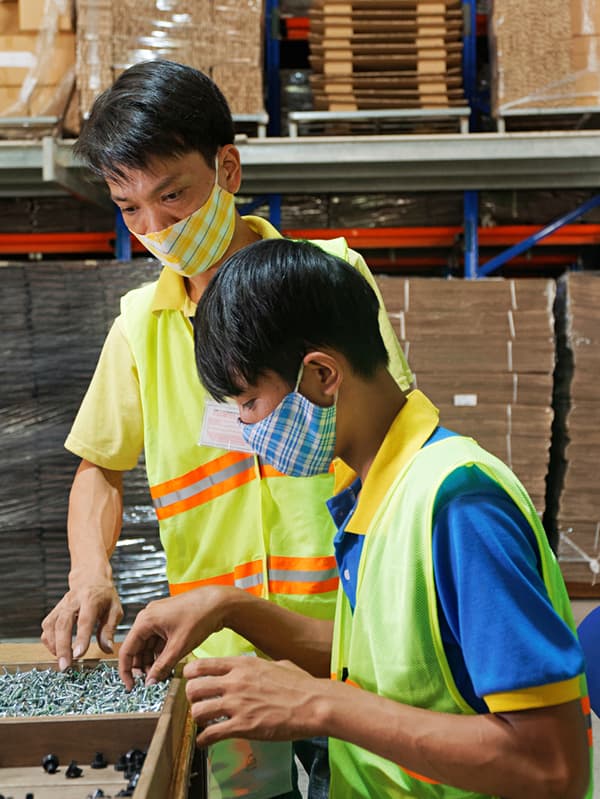 Dous traballadores de IKEA con chalecos de seguridade amarelos e máscaras examinando parafusos nunha fábrica.