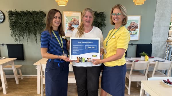 Drei Frauen stehen im IKEA Restaurant nebeneinander, im Hintergrund sind Holztische, Stühle, Pflanzen und Wanddeko zu sehen. Zwei der Frauen tragen IKEA-Mitarbeiterkleidung und überreichen einen Spendenscheck über 5.000 Euro an die Vertreterin des Kinderschutzbundes Siegen-Wittgenstein, die Freizeitkleidung trägt.