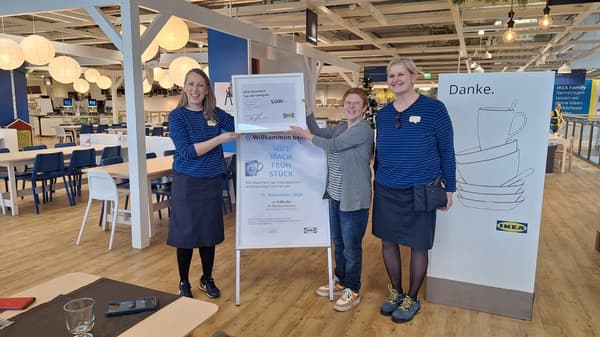 Drei Frauen stehen im Restaurantbereich eines IKEA-Marktes und halten gemeinsam einen gerahmten Spendenscheck über 5.000 Euro. Zwei der Frauen tragen blaue IKEA-Uniformen mit Streifen, während die dritte Person in einer grauen Strickjacke gekleidet ist. Im Hintergrund steht ein Plakat mit der Aufschrift „MUT MACH FRÜHSTÜCK“, das auf eine Veranstaltung für Frühcheneltern hinweist.  