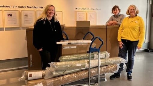 Drei Frauen stehen in einem IKEA-Markt. Zwei von ihnen tragen Freizeitkleidung, eine trägt eine gelbe IKEA-Mitarbeiterjacke. Vor ihnen stehen Transportwagen mit aufgerollten Teppichen und Kartons mit Möbelspenden.  