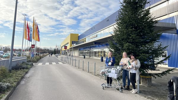 Drei Frauen stehen mit einem Einkaufswagen voller Spenden vor einem großen IKEA-Gebäude. Der Himmel ist blau mit einigen Wolken, mehrere Landesflaggen sind auf Fahnenmasten zu sehen. Im Hintergrund erstreckt sich eine breite Straße mit weiteren Personen und Autos.  